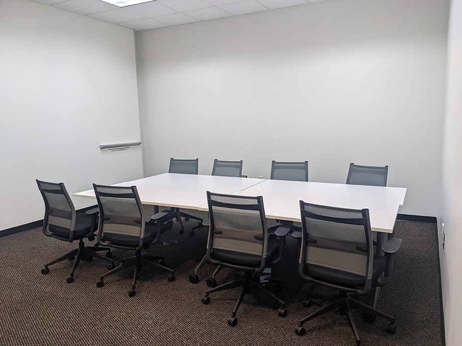Small meeting room with a modular white table and mesh office chairs arranged for group work, set against plain walls.