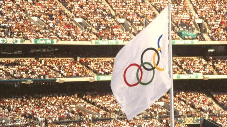 Olympic flag with the five rings flies in front of a packed stadium filled with spectators during a large sporting event.