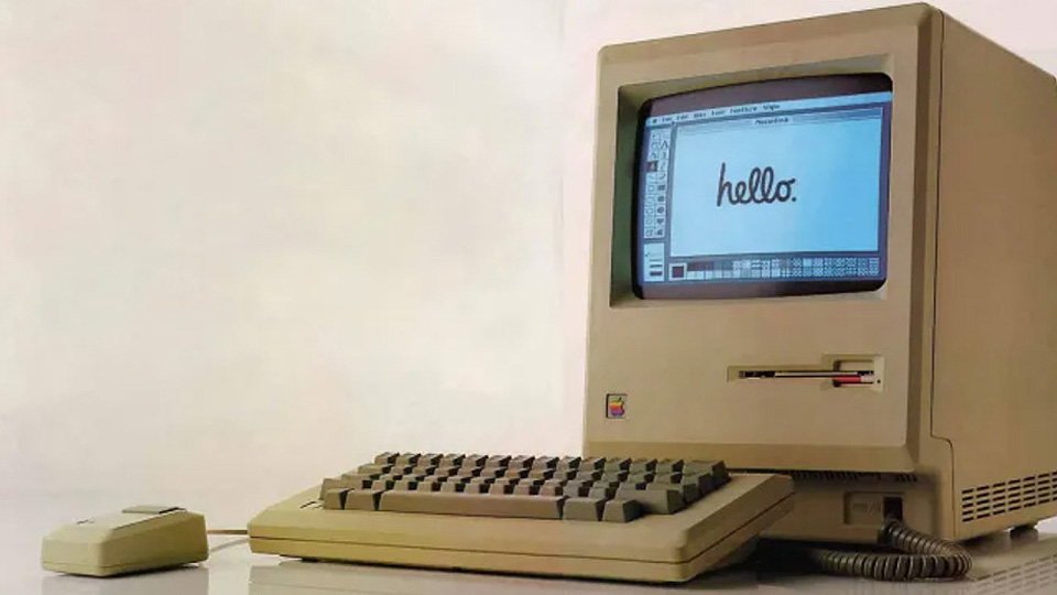 Vintage Apple Macintosh computer with keyboard and mouse displaying a handwritten “hello.” message on its monochrome screen.