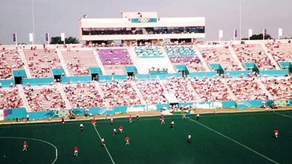 Spectators fill a large stadium while athletes compete on a field during a sporting event viewed from the stands.