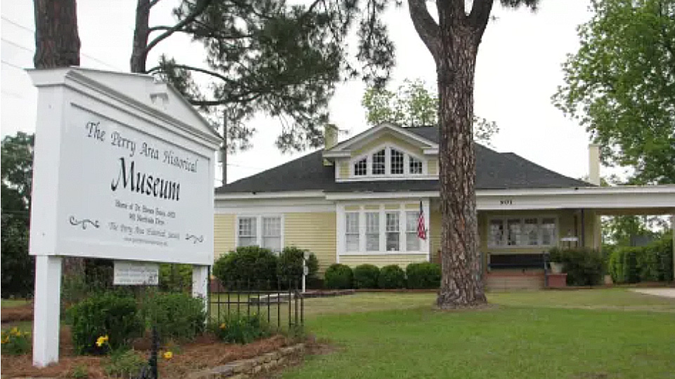 White museum sign stands in front of a historic house surrounded by trees and a lawn at the Perry Area Historical Museum.