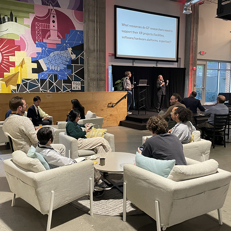 Event space with attendees seated in lounge chairs facing a stage, where two speakers present beside a large screen displaying a question about XR research resources. Colorful mural and “TSQ ATL” text visible on the wall.