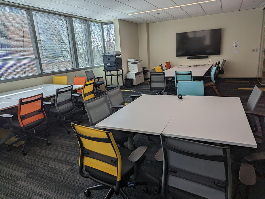 Flexible classroom or meeting space with grouped tables, mesh office chairs in various colors, large wall-mounted display, printer, and windows providing natural light.