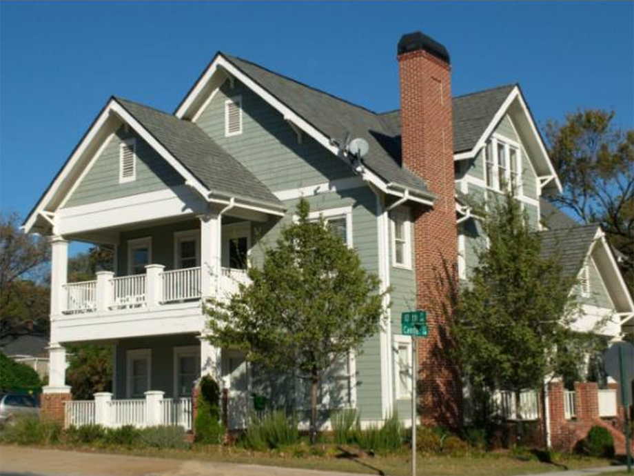 Two-story light green house with white trim, a covered front porch and balcony, a brick chimney, and surrounding trees along a street corner.