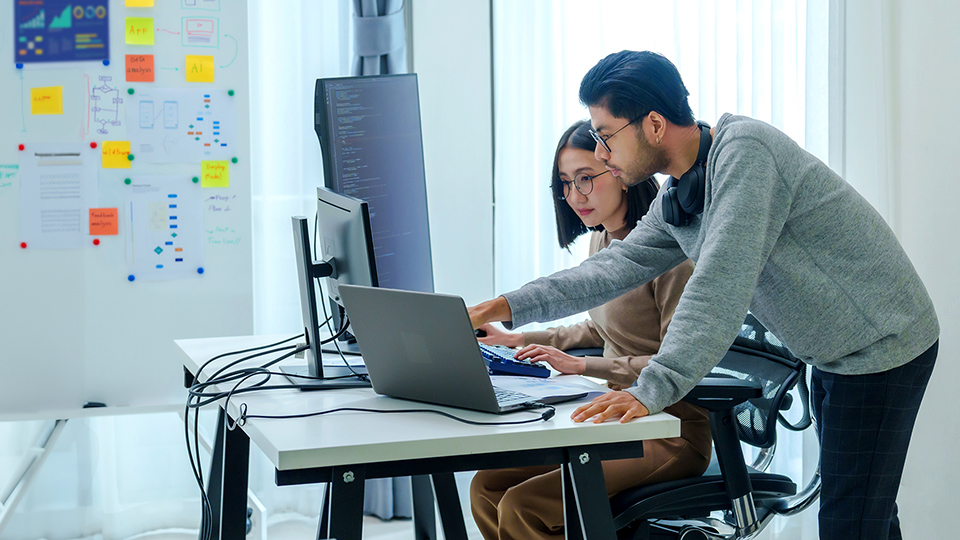 Two people collaborate at a desk with laptops and monitors, reviewing code in a bright office with notes on the wall.