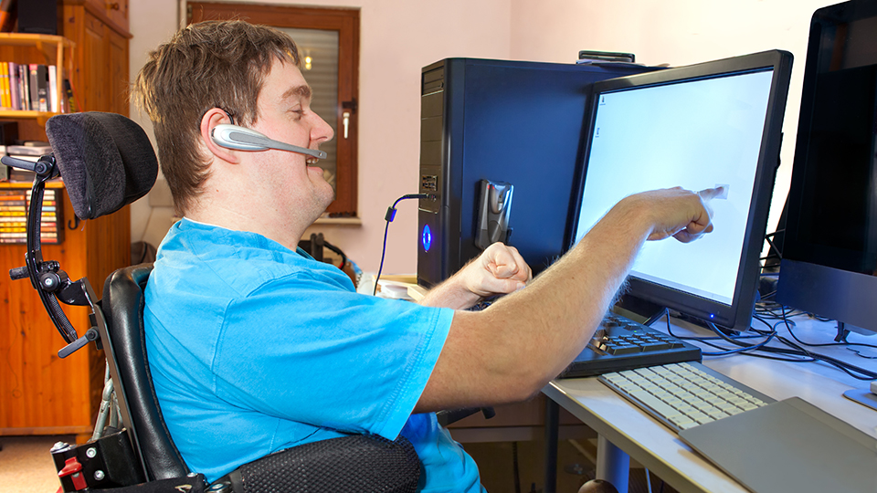 Person using a wheelchair and headset while interacting with a computer workstation equipped with multiple monitors and assistive technology.
