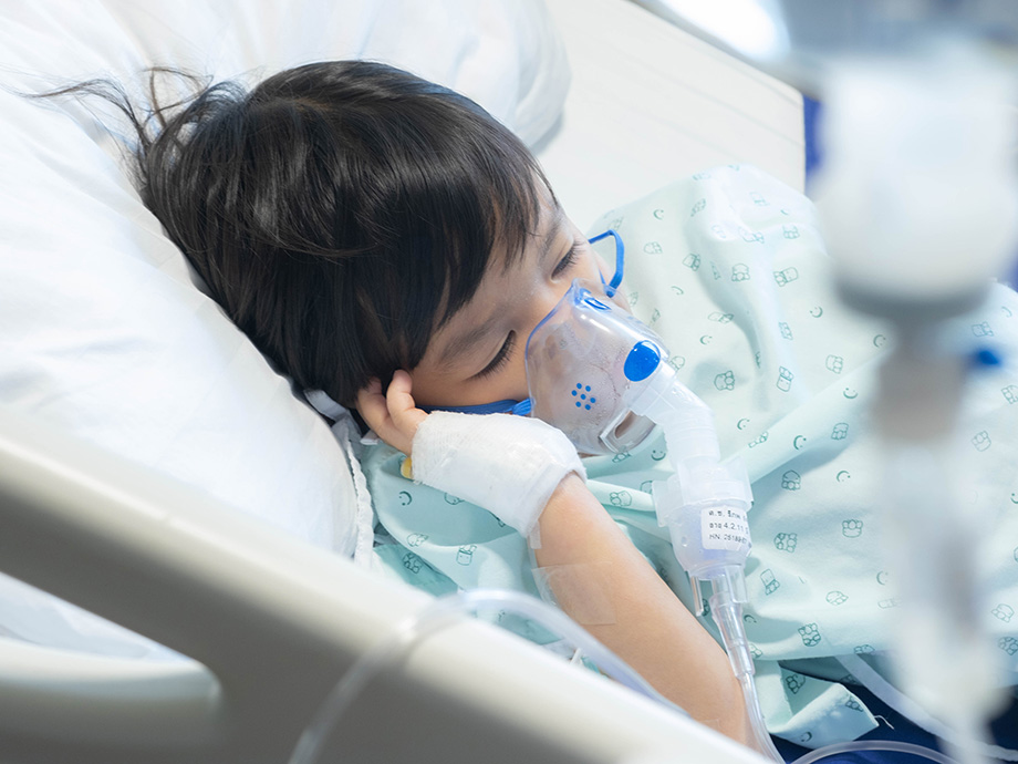 Child lying in a hospital bed wearing an oxygen mask, with an IV line in place and medical equipment surrounding the bedside.