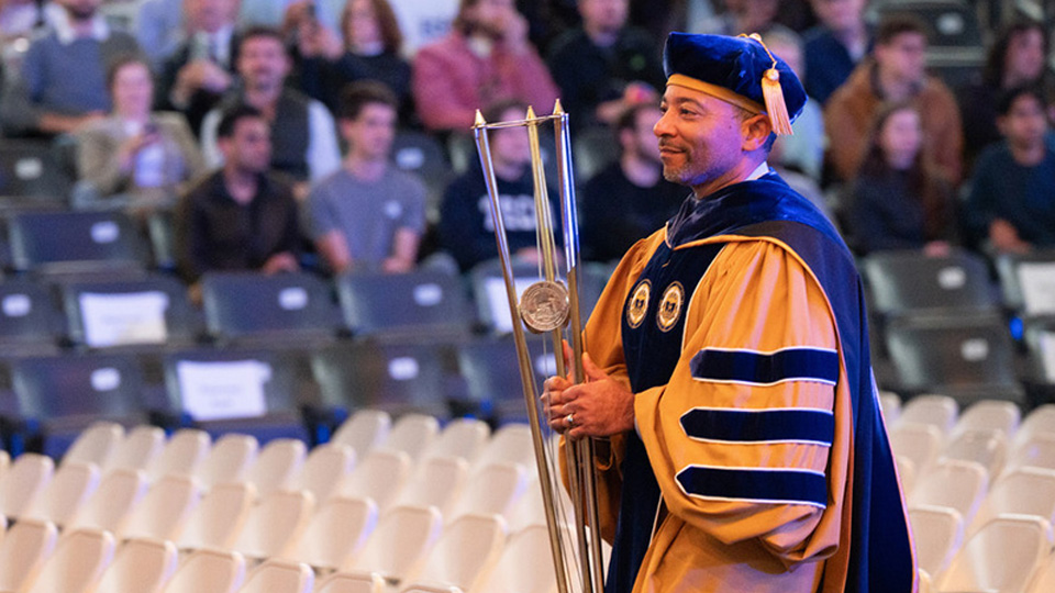 Academic official in gold and blue regalia carries a ceremonial mace in an arena with blurred audience seating in the background.