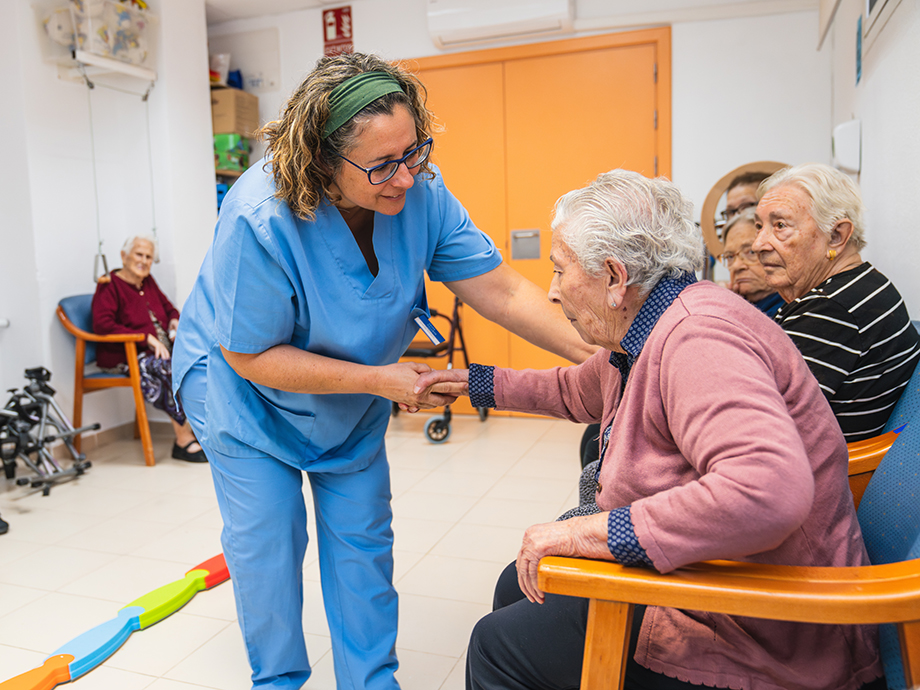 Caregiver in a clinical room gently assists an older adult seated in a chair, while other individuals sit nearby with mobility aids visible in the background.