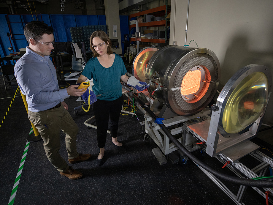 Two people wearing protective eyewear stand beside a large cylindrical testing chamber, one holding a device while the other adjusts equipment connected to the glowing interior.