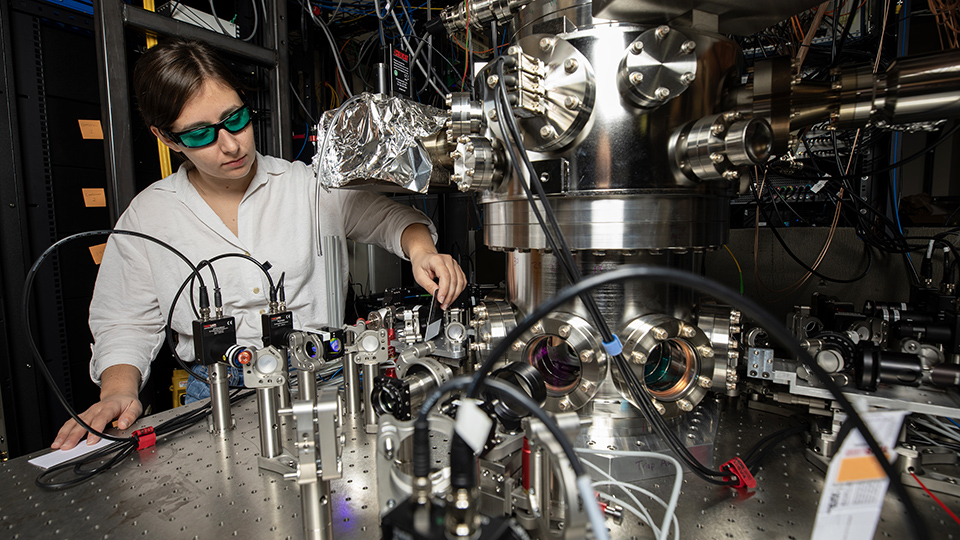 Researcher adjusts complex lab equipment with lenses, mirrors, and wiring in a high-tech laboratory setup.
