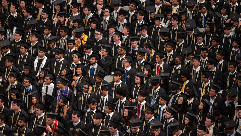 A dense crowd of graduates seated indoors, all wearing black caps and gowns with gold tassels and cords. Rows of attendees fill the frame, creating a repeating pattern as individuals adjust caps, look around, or hold programs during the ceremony.