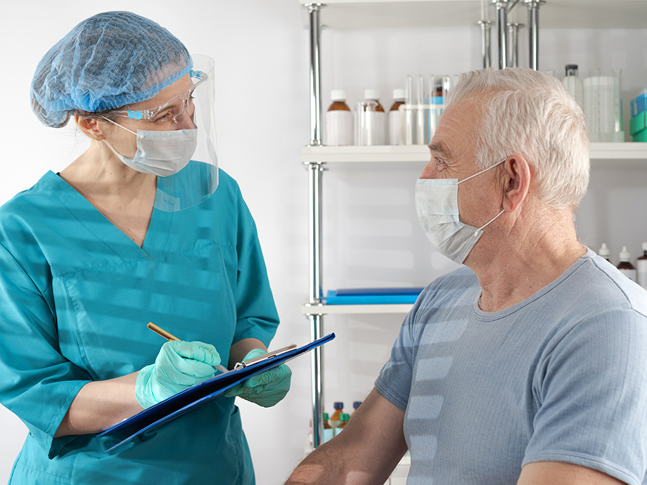 Healthcare professional wearing protective gear writes on a clipboard while speaking with a seated patient in a clinical room.