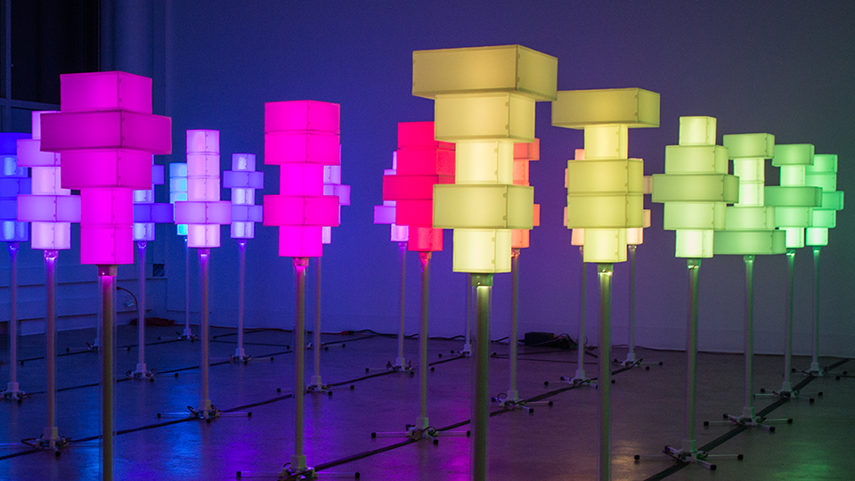 Colorful illuminated geometric light sculptures on stands arranged in rows inside a dark gallery space.