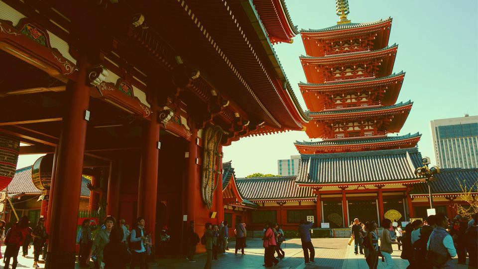 Visitors walk through the grounds of a traditional Japanese temple complex featuring red wooden buildings and a five-story pagoda.