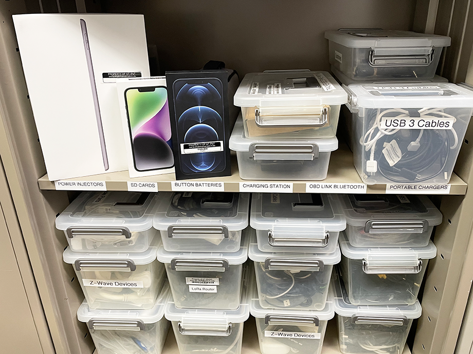 Shelves filled with labeled plastic storage bins containing cables, devices, and electronics, alongside boxed tablets and phones.