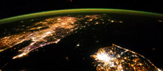 Nighttime view of Earth from space, showing illuminated city lights along coastlines beneath the planet’s curved horizon.