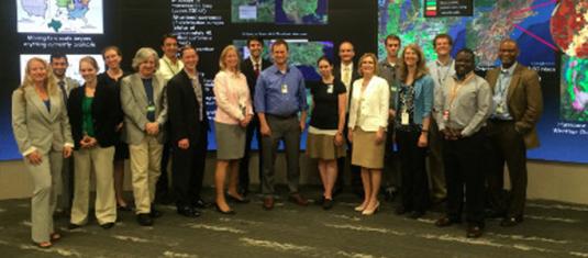 Group photo in a research operations center, with large digital displays showing maps, data visualizations, and imagery.