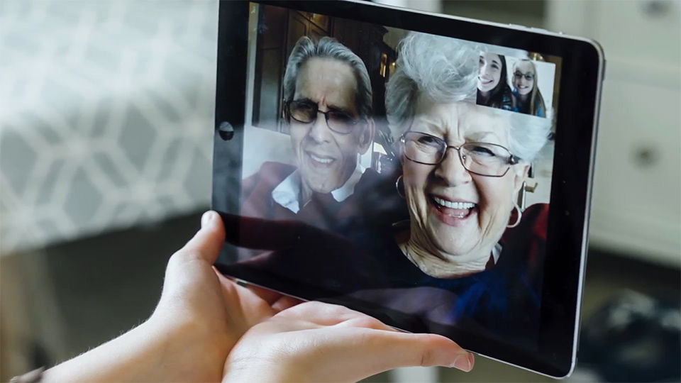 Tablet showing a video call with two older adults, with a smaller inset of another participant, held in someone’s hands.