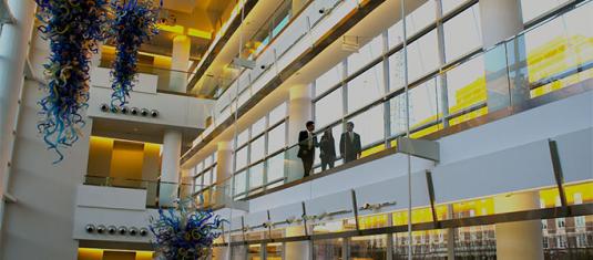 Interior atrium with glass walls, suspended art installations, and people walking along an upper-level corridor.