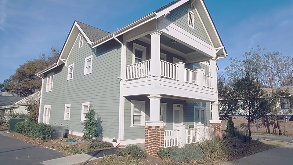Two-story light green house with white trim, front porch, and balcony, set along a paved driveway in a residential area.