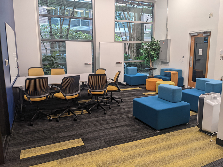 Collaborative workspace with tables, rolling chairs, whiteboards, and colorful lounge seating near large windows bringing in natural light.