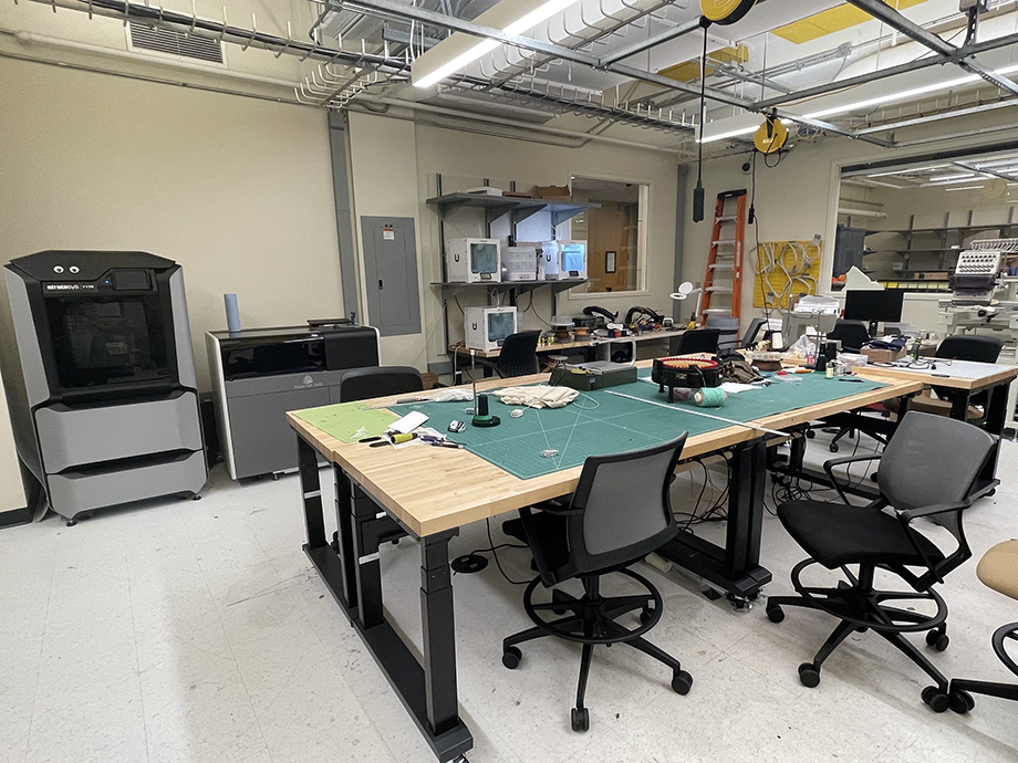 Maker space with worktables, chairs, tools, and 3D printers, set in a bright lab with exposed ceilings and organized equipment.