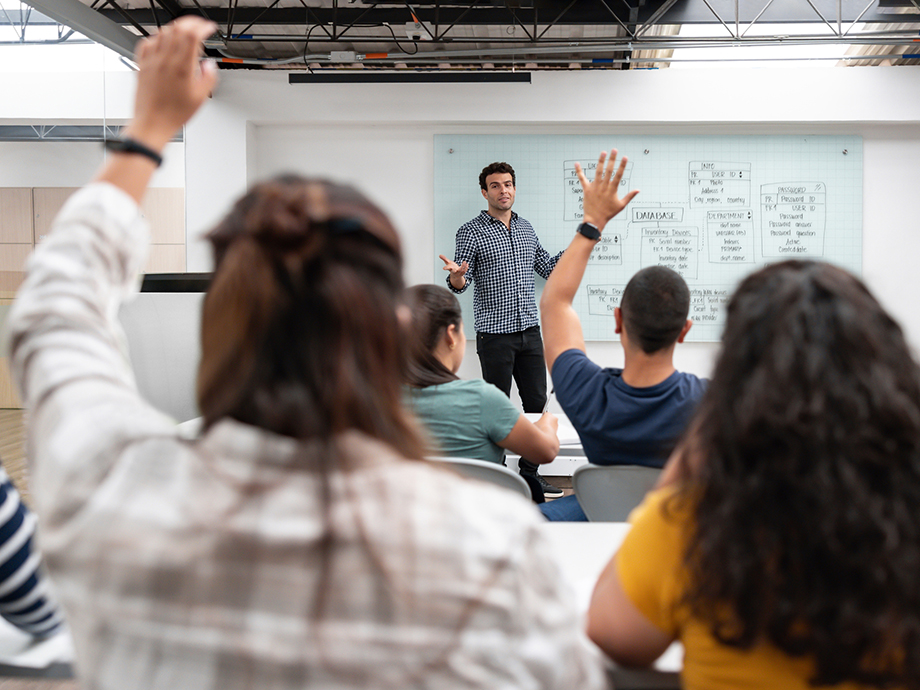 Instructor stands at the front of a classroom near a whiteboard filled with diagrams, while several seated students raise their hands.