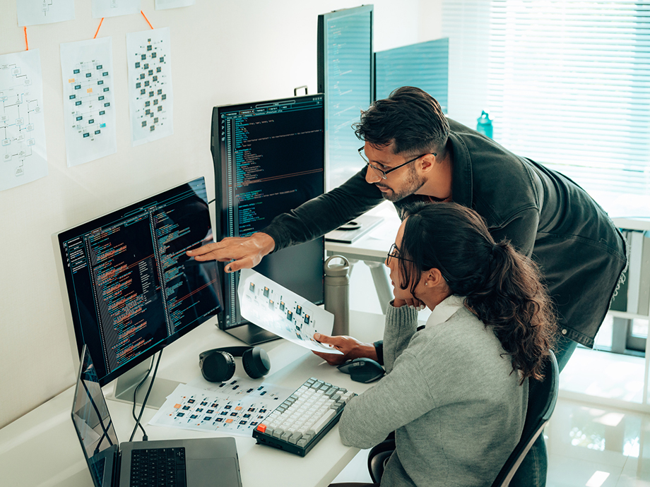 Two people collaborate at a desk, reviewing code on dual monitors while one points at the screen and the other holds a document.
