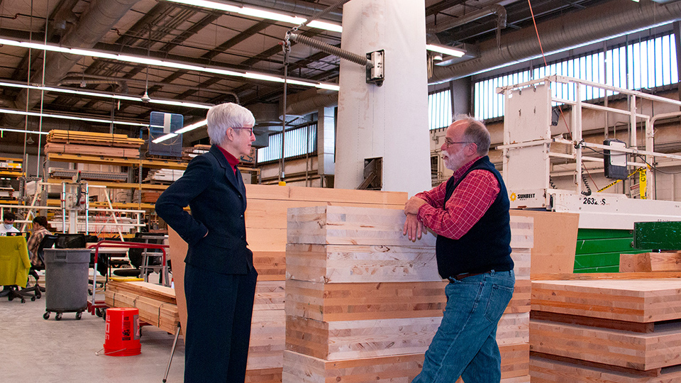 Two people stand and talk in a large woodworking and manufacturing facility surrounded by stacked butcher-block wood panels, industrial equipment, and workshop materials.