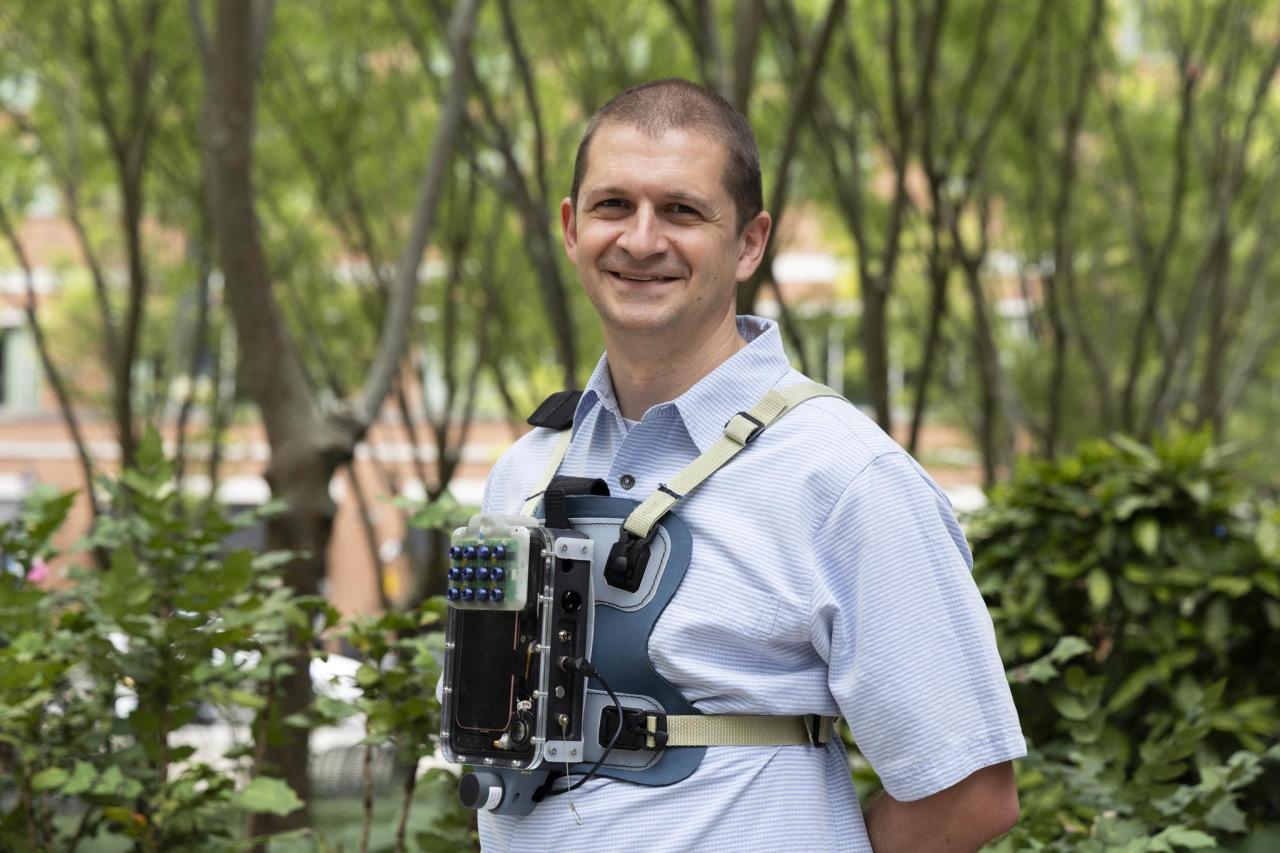 Person standing outdoors in a tree-filled area, wearing a strapped chest-mounted electronic device with buttons and wiring, positioned in front of leafy greenery and blurred buildings in the background.