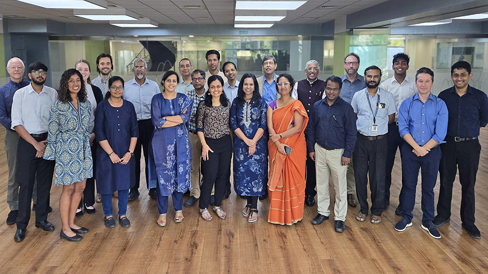 Group of people standing together in a bright office space, arranged in rows on a wooden floor for a group photo.