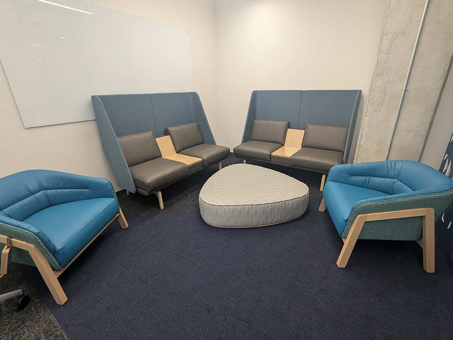 Small lounge-style collaboration area with cushioned blue chairs, two high-back benches, and a round ottoman arranged around a central space near a whiteboard.