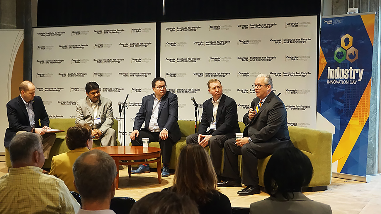 Panel discussion on a stage with five speakers seated on a couch and chairs, speaking into microphones before an audience, with a Georgia Tech–branded backdrop and an “Industry Innovation Day” banner.