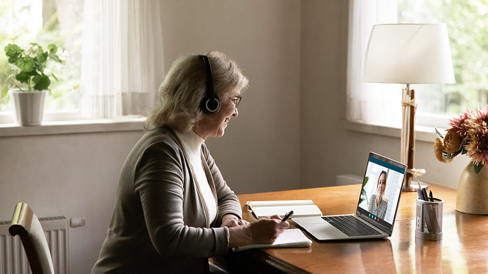 Person wearing headphones sits at a desk, taking notes while video chatting on a laptop in a bright room.