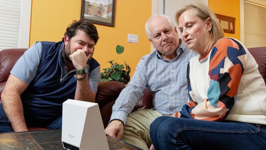 An older couple sitting on a couch as a man helps them use Amazon's Alexa