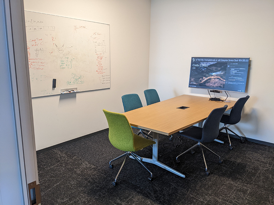 Small meeting room with a wooden table, rolling chairs, a large whiteboard with notes, and a monitor displaying information on the wall.