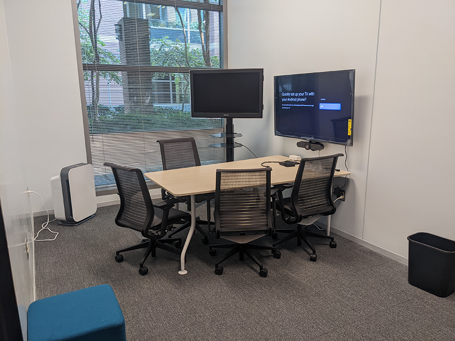 Compact meeting room with a small table, mesh chairs, dual monitors, and a wall-mounted display for presentations.